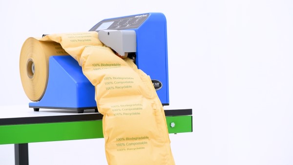 Packaging machine dispensing brown paper cushions labeled '100% Biodegradable, 100% Compostable, 100% Recyclable' on a table.