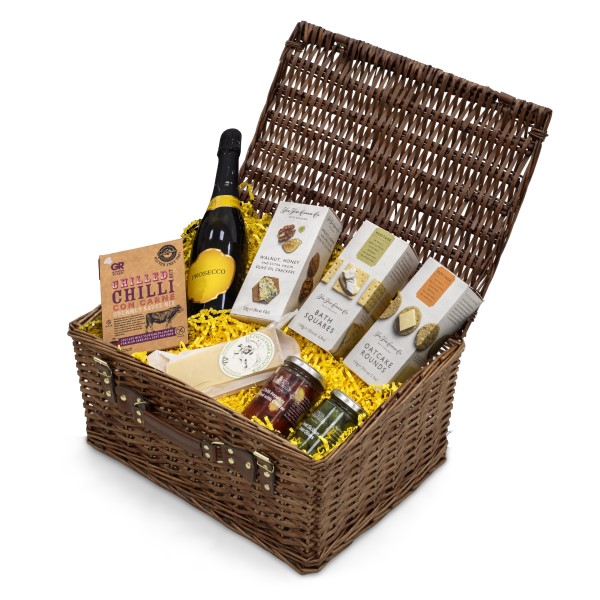 Wicker hamper with a layer of shredded paper and filled with prosecco, snacks, crackers, cheese, and a jar of preserves.